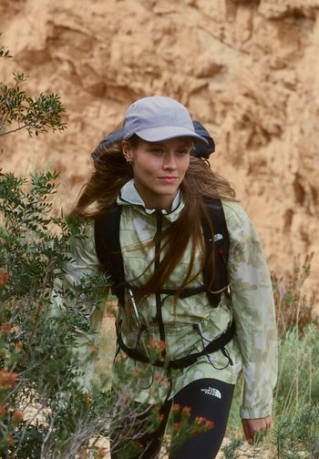 Frau mit Mütze, Tarnjacke und Rucksack wandert durch grüne Büsche, im Hintergrund felsiges Gelände.