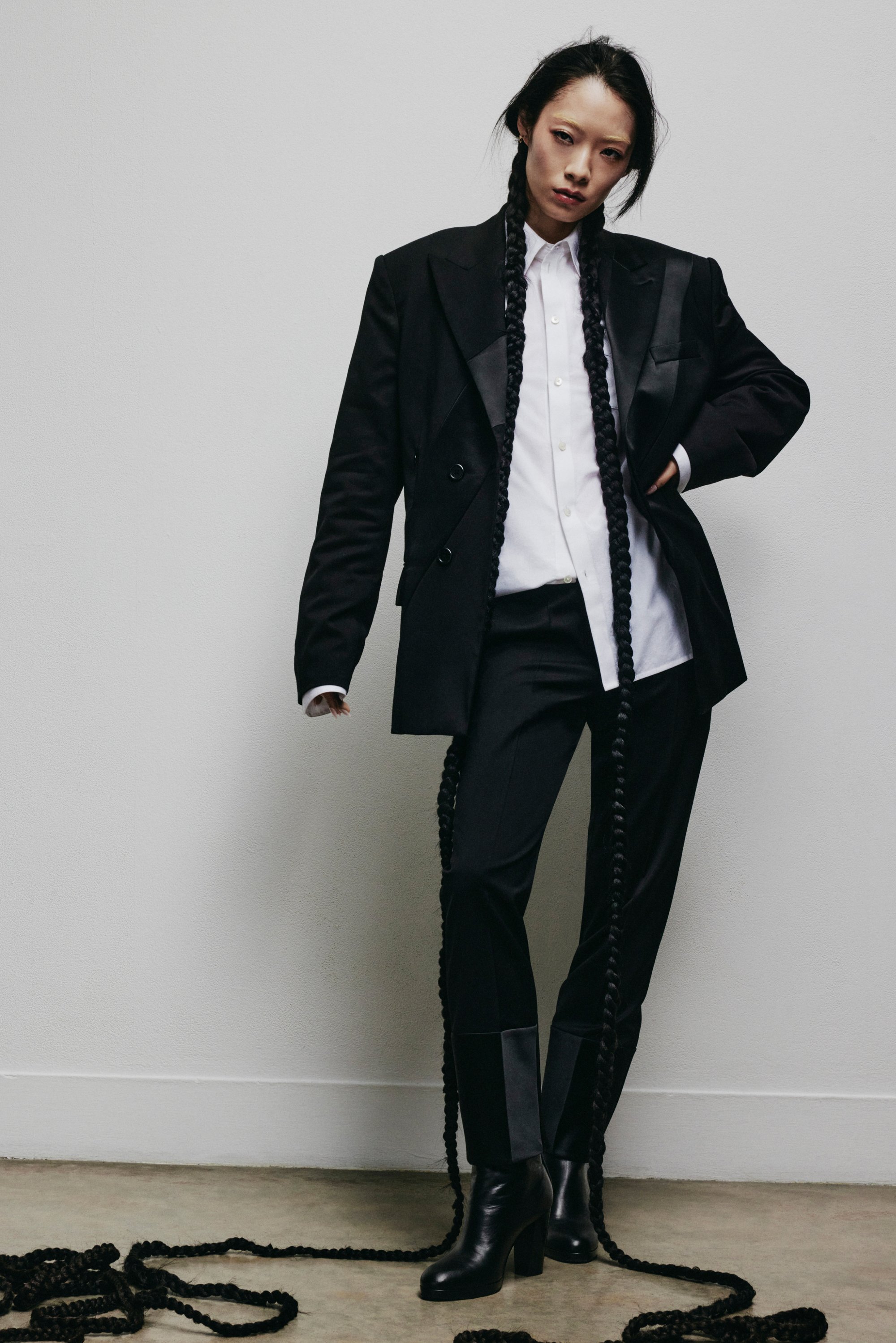 Fashion model in a tailored black suit with a white shirt, sporting long braided hair cascading around her. She stands confidently against a neutral background.
