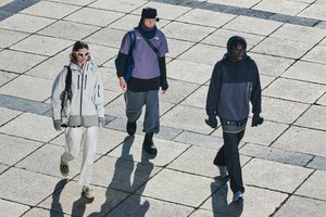 Three individuals walk on a stone pavement, wearing fashionable outdoor clothing. Sunlight casts shadows, highlighting their unique styles.