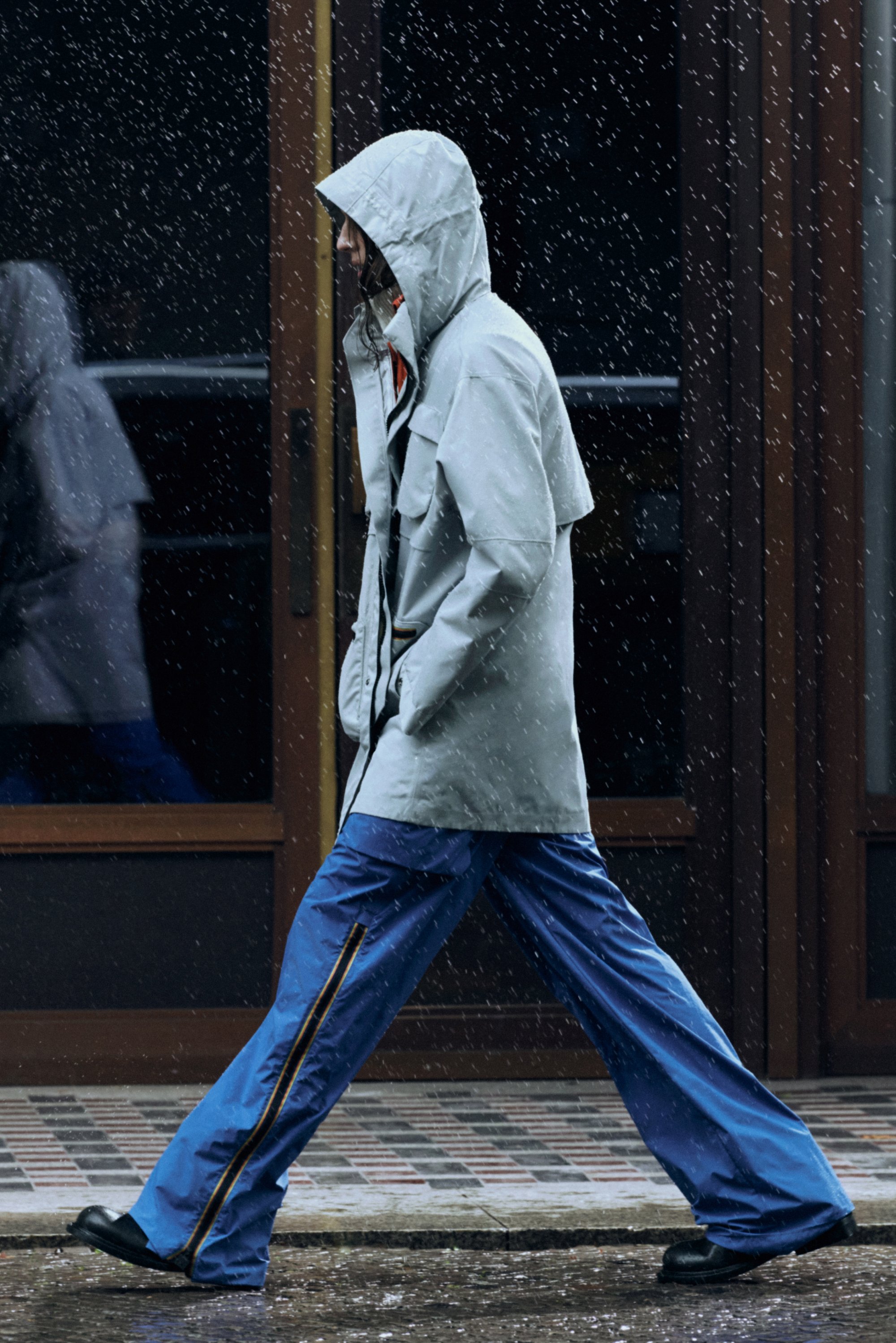 Helle graue, wasserabweisende Jacke mit Kapuze und zwei Fronttaschen, kombiniert mit lockeren blauen Hosen mit seitlichen Streifen.