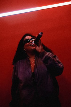 Person with curly hair wearing sunglasses, singing into a microphone against a red background, illuminated by diagonal white light above.