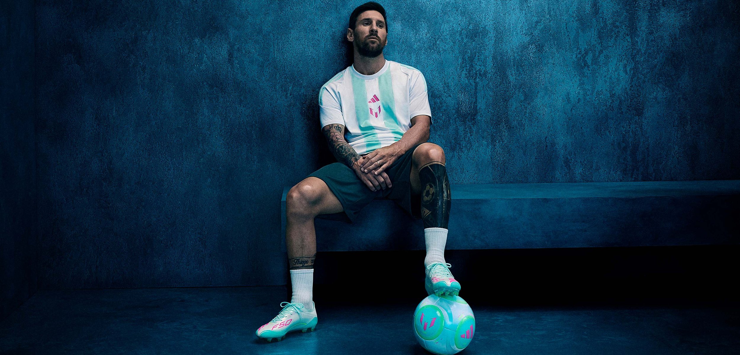 Homme avec des tatouages assis sur un banc bleu, reposant un pied sur un ballon de football aux couleurs pastel, devant un mur texturé bleu foncé.