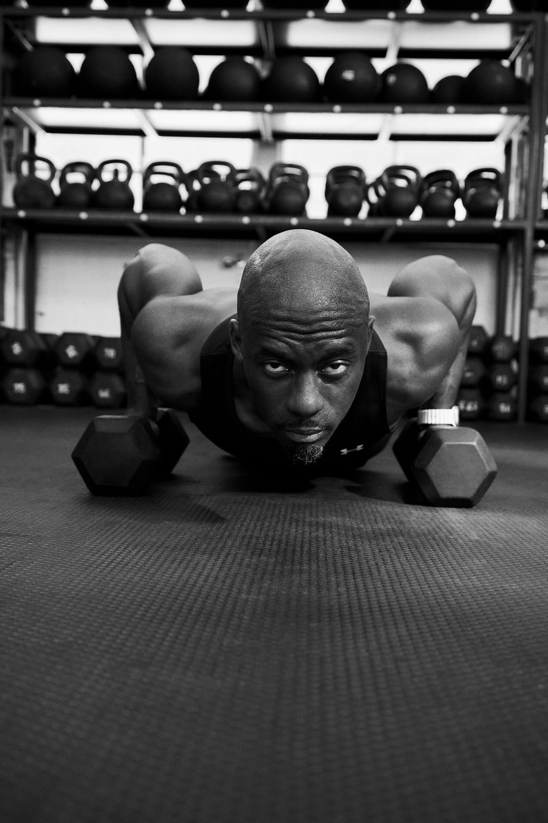 Svalnatá osoba vykonáva kľuky na telocvičnom koberci, čelom k fotografovi, s jednoručnými činkami vedľa seba a s činkami v pozadí.