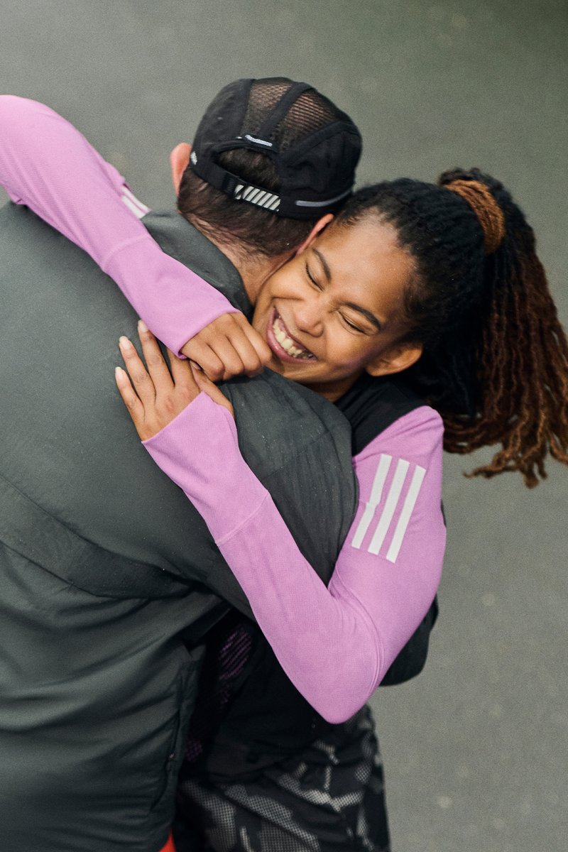 A woman with dreadlocks happily hugs a man from behind, both smiling. She wears a pink long-sleeve shirt, while he has on a grey jacket.