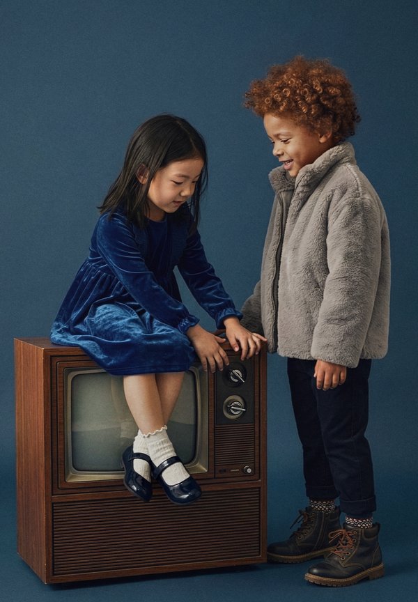 Une fille en robe bleue est assise sur une télévision vintage tandis qu'un garçon en veste grise se tient debout en souriant, tenant sa main, sur un fond sombre.