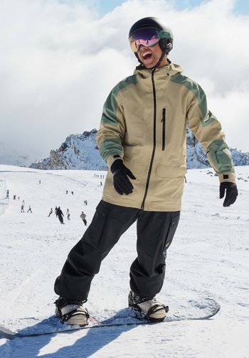 Chaqueta de snowboard beige y verde con pantalones negros, guantes negros y botas de esquí blancas en un paisaje nevado, mostrando deportes de invierno activos.