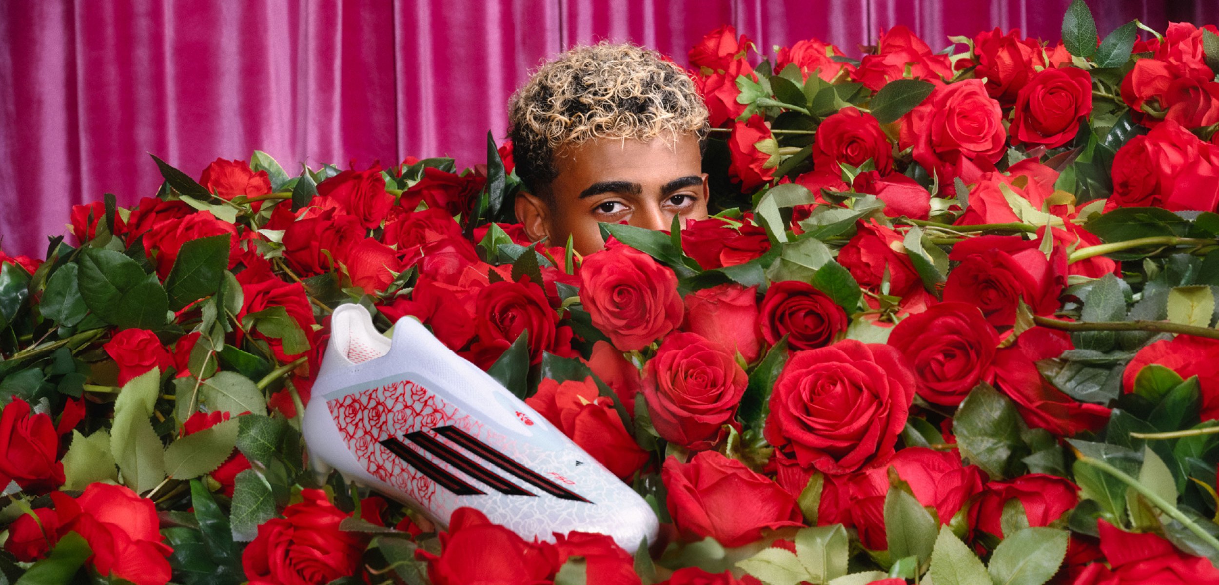 Personne aux cheveux blonds bouclés partiellement cachée parmi des roses rouges, chaussure de sport blanche et rouge bien visible au-dessus des roses.