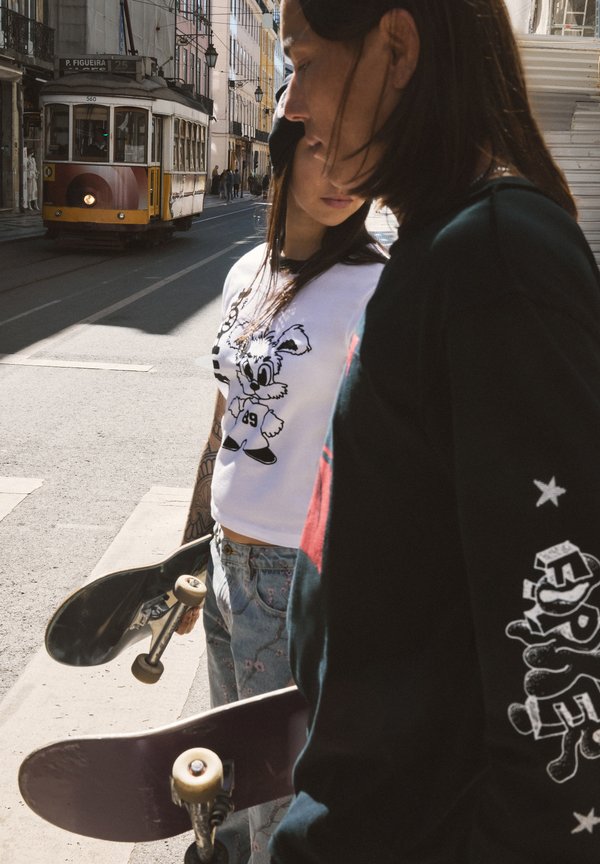 Two young people holding skateboards stand on a city street with a tram approaching in the background.