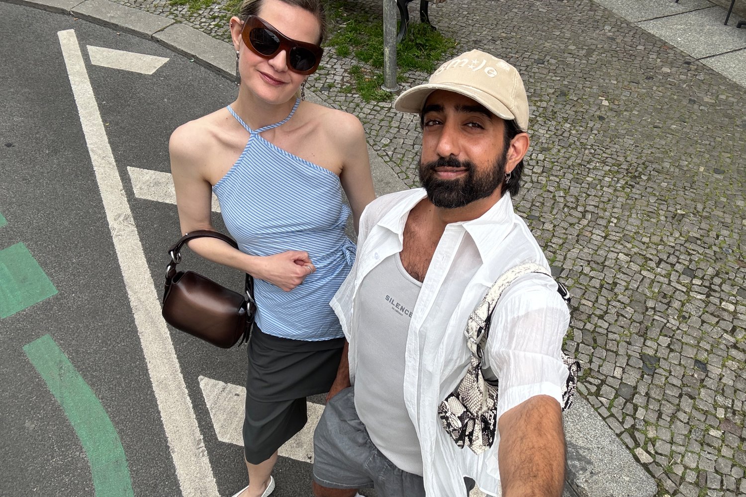 Due amici posano per un selfie su una strada acciottolata. Uno indossa una maglietta a righe blu e porta una borsa marrone; l'altro è vestito con un cappello e un abbigliamento chiaro.