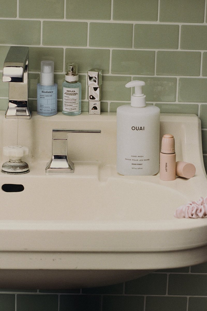 Waschbecken im Badezimmer mit Chromarmatur, Biodance- und Replica-Serumflaschen, Ouai-Handseifenflasche, Inglot-Make-up-Stift und rosa Haargummi an der gefliesten Wand.