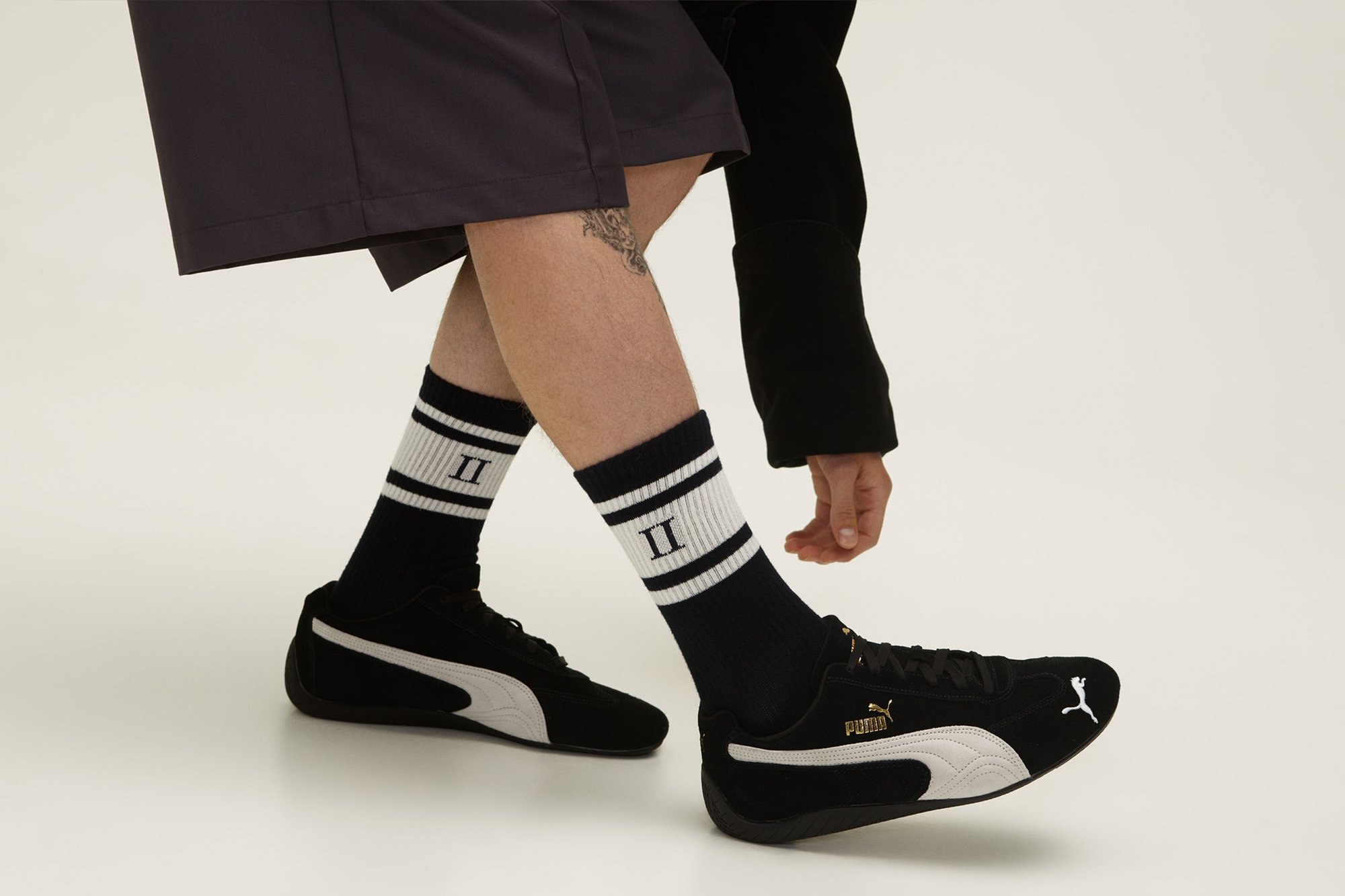 Zapatillas deportivas de ante negro con detalles en blanco, combinadas con calcetines a rayas en azul marino y blanco, llevadas con pantalones cortos oscuros. Detalle del logo dorado notable.