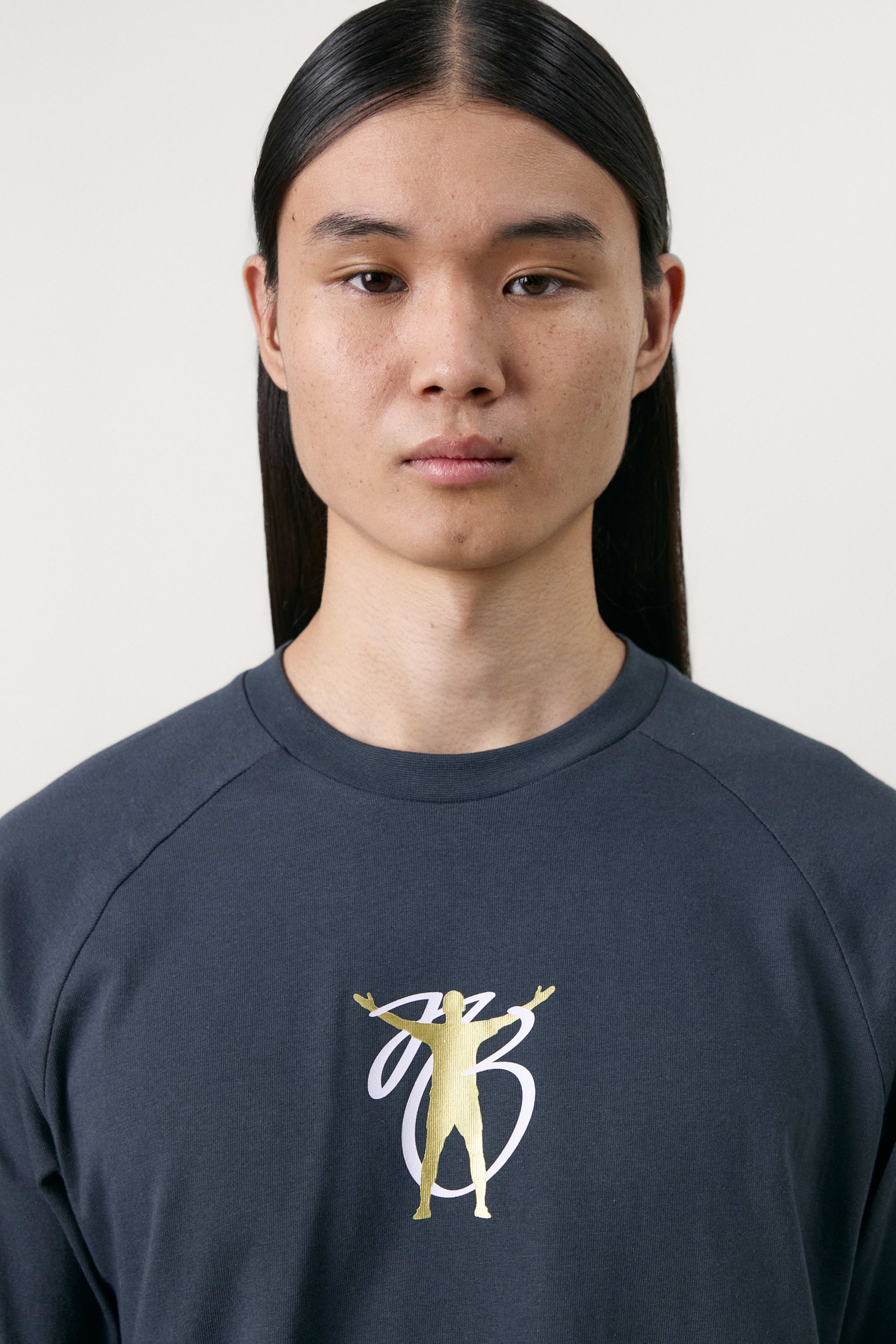 Young person with long hair in a dark sweatshirt featuring a gold and white graphic of a figure with arms raised against a light background.