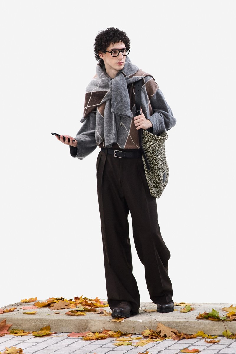 Pullover mit grauem und braunem Rautenmuster, dicker Schal übergeworfen, lockere braune Hose, schwarze Schuhe, hält ein Handy, trägt eine strukturierte Tasche.