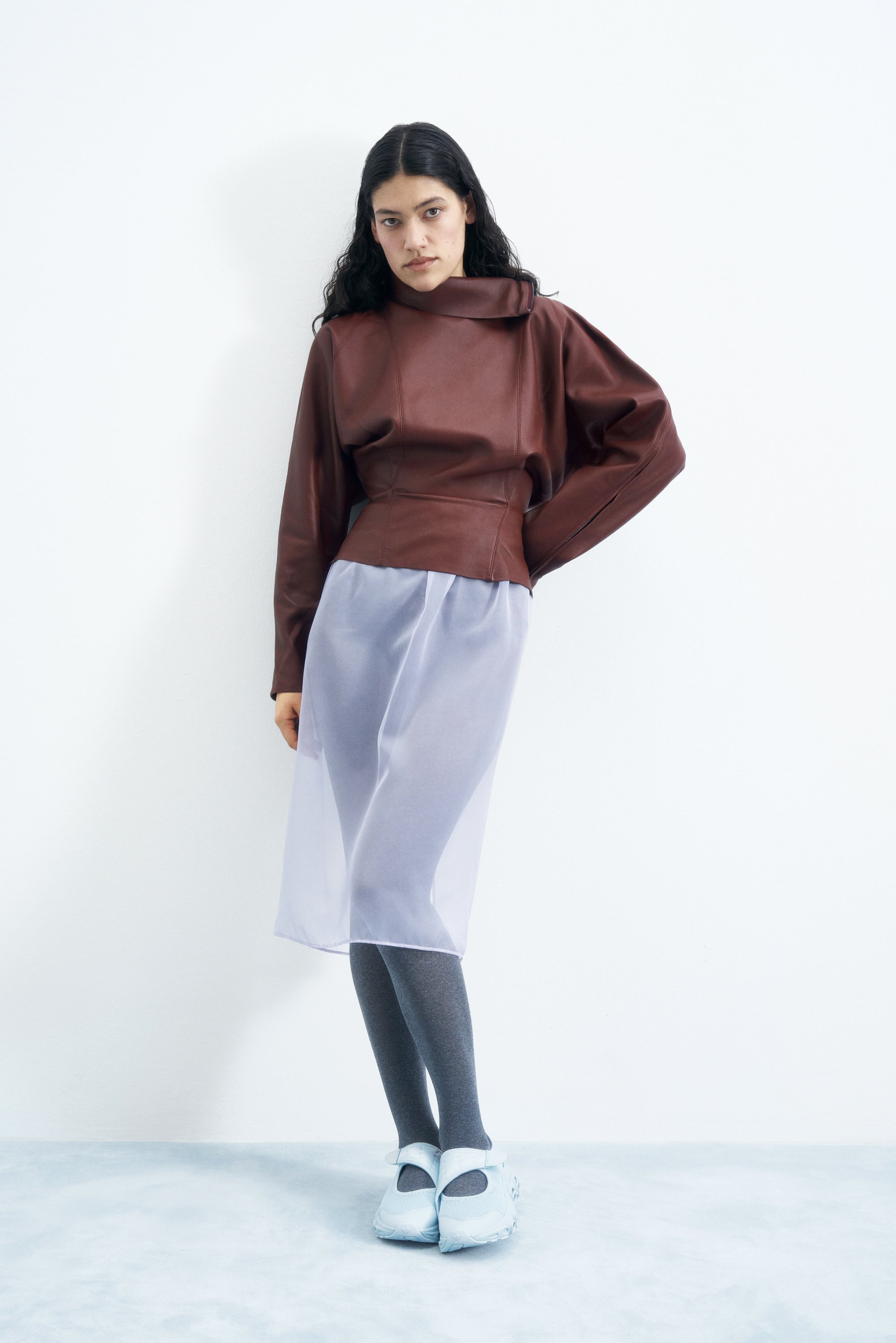 Una persona con cabello largo y oscuro lleva un top de cuero marrón, una falda transparente y zapatos de color azul claro, de pie contra un fondo blanco liso.