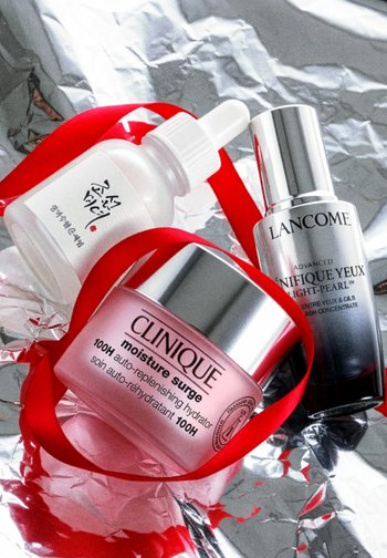 Barattolo di Clinique Moisture Surge, flacone di siero Lancome Advanced Génifique Yeux e flacone con contagocce di prodotti skincare coreani con nastro rosso su foglio di alluminio argentato.