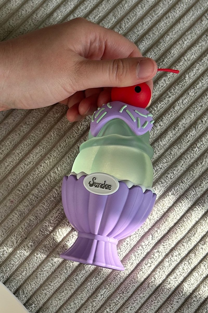 Hand hält ein lila-grünes Plastik-Eisbecher-Spielzeug mit einer roten Kirsche oben drauf und einem "Sundae"-Etikett am Becher.