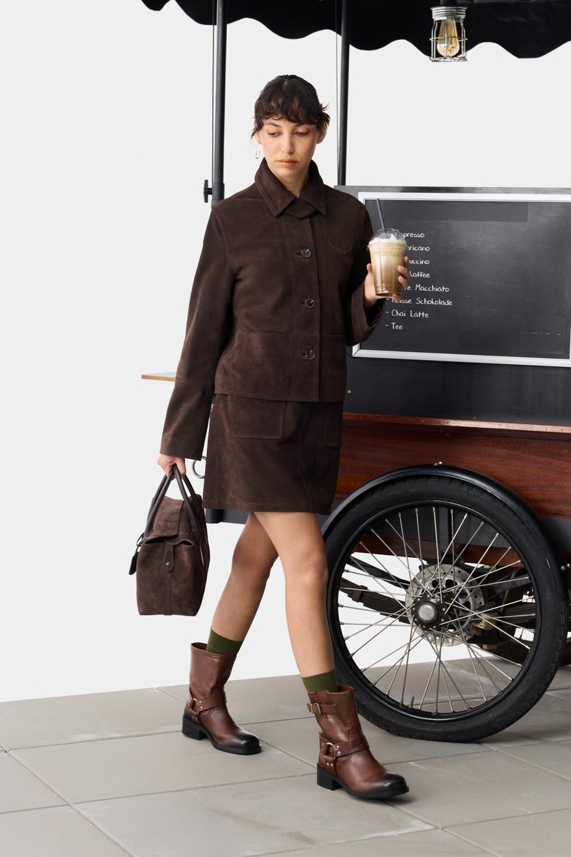 Braunes Wildleder-Jacken- und Rock-Set, braune Lederstiefel mit Schnallen, grüne Socken, hält eine braune Tasche und steht neben einem Kaffeewagen.