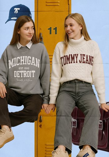 Two young women sit on a yellow locker, holding hands. One wears a gray Michigan sweatshirt, the other a cream cable-knit sweater.