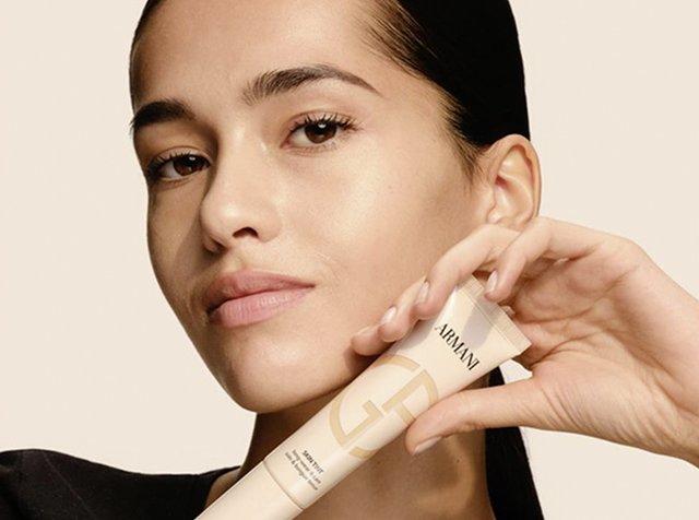 Tubo beige de productos de cuidado de la piel de Armani sostenido por una persona con piel suave. El tubo presenta un diseño minimalista con un logo prominente y un acabado mate.