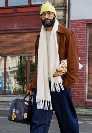 Brown suede jacket over a cream jumper, blue wide-leg jeans, white fringed scarf, yellow ribbed beanie, holding a black bag with patches and bread.