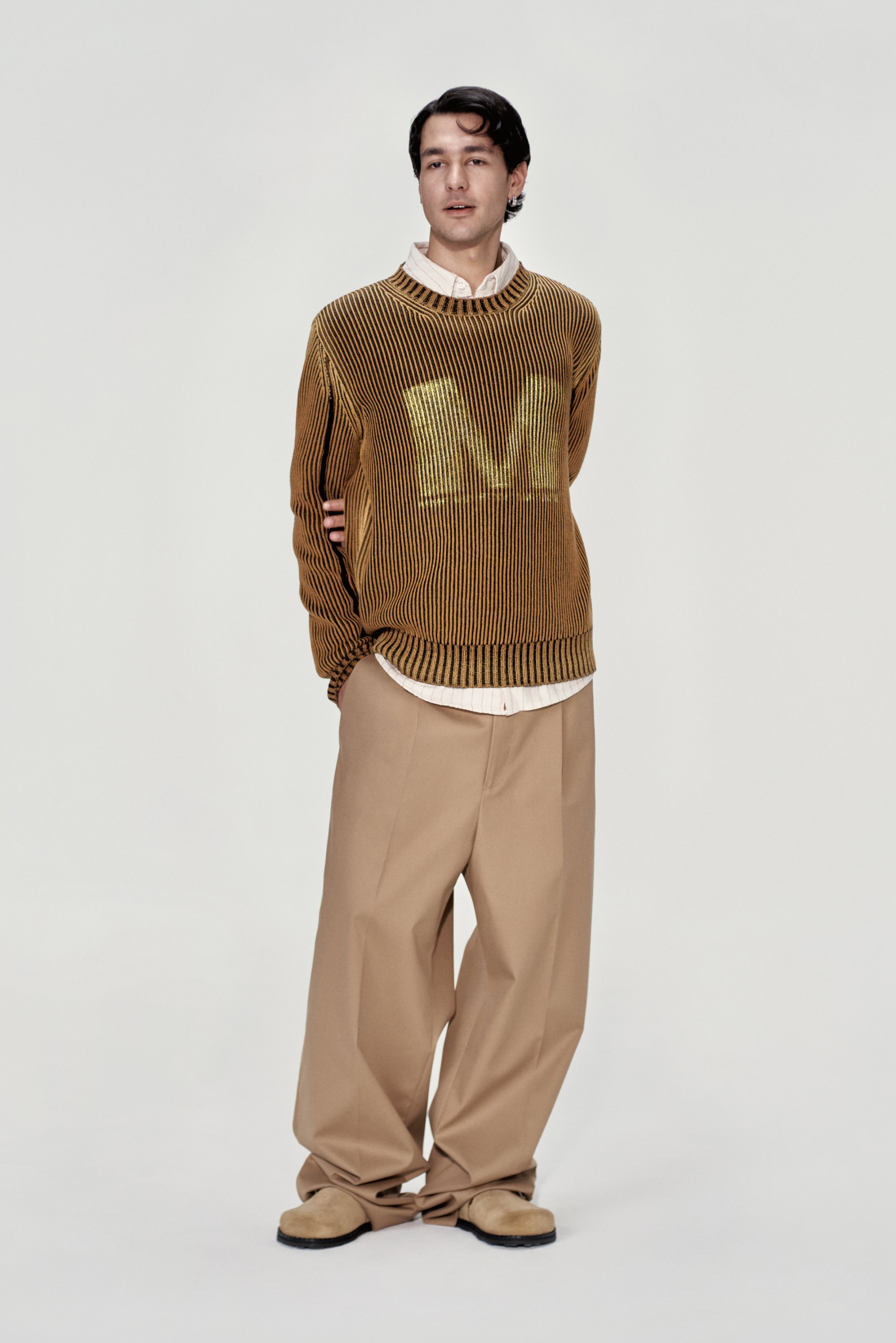 Maglione a coste marrone con un grande logo "M", indossato sopra una camicia button-up beige chiaro, abbinato a pantaloni larghi color sabbia e scarpe marroni.