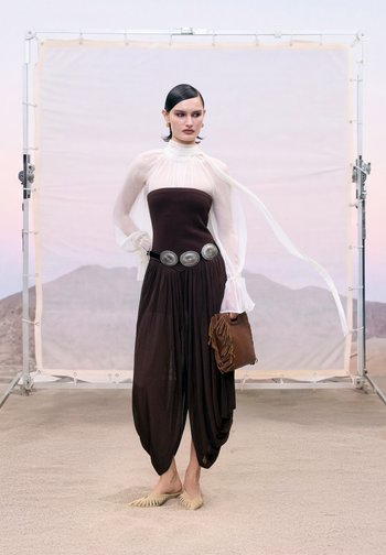 Femme portant un chemisier blanc transparent et un pantalon harem noir sans bretelles avec une ceinture argentée, tenant une pochette marron à franges, debout sur un sol sablonneux.