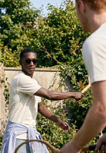 Dos hombres jugando al tenis al aire libre, uno con gafas de sol y una camisa clara, balanceando una raqueta de madera.