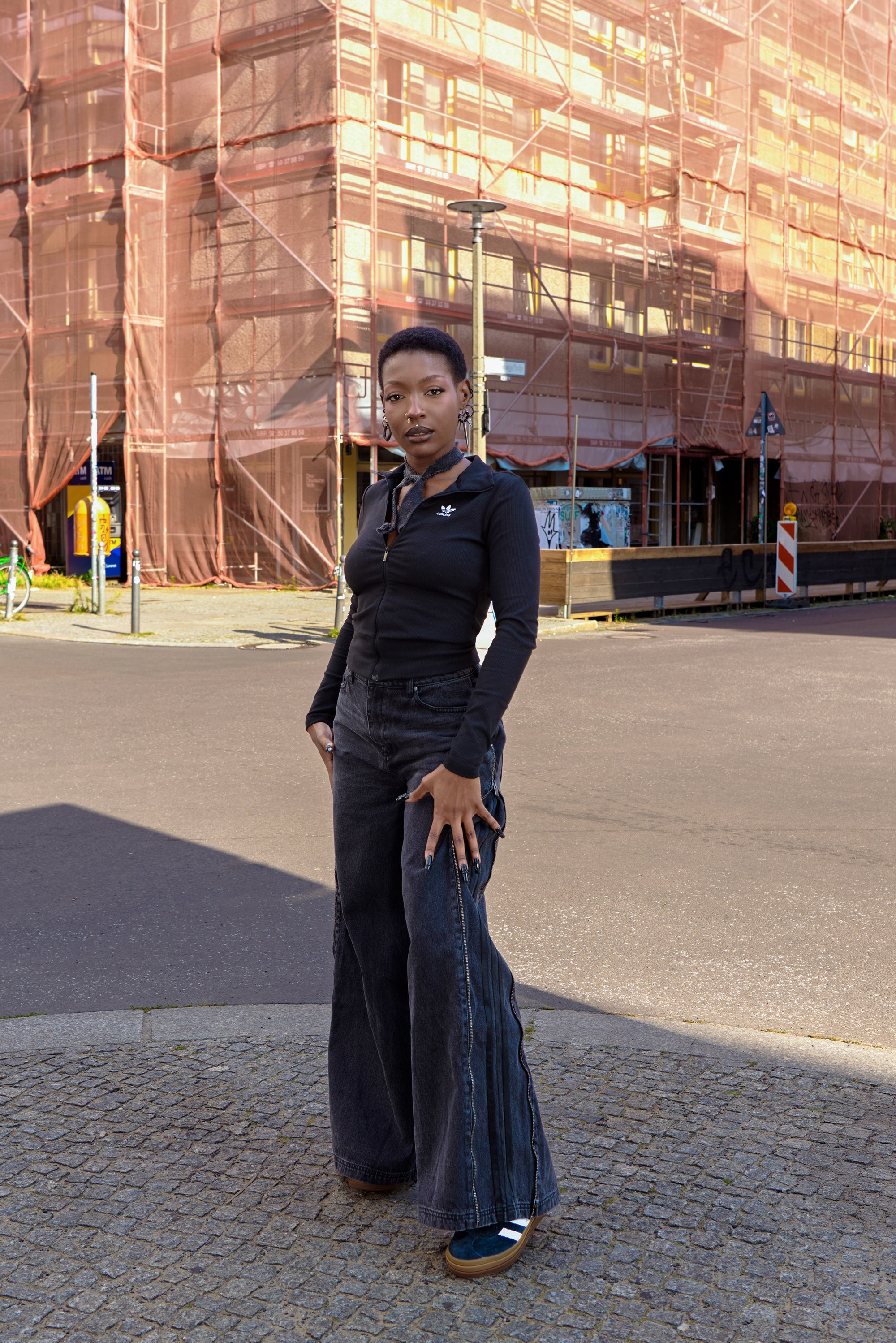 Person wearing black zip-up top and wide-leg jeans stands on cobblestone street near building covered in scaffolding and netting.