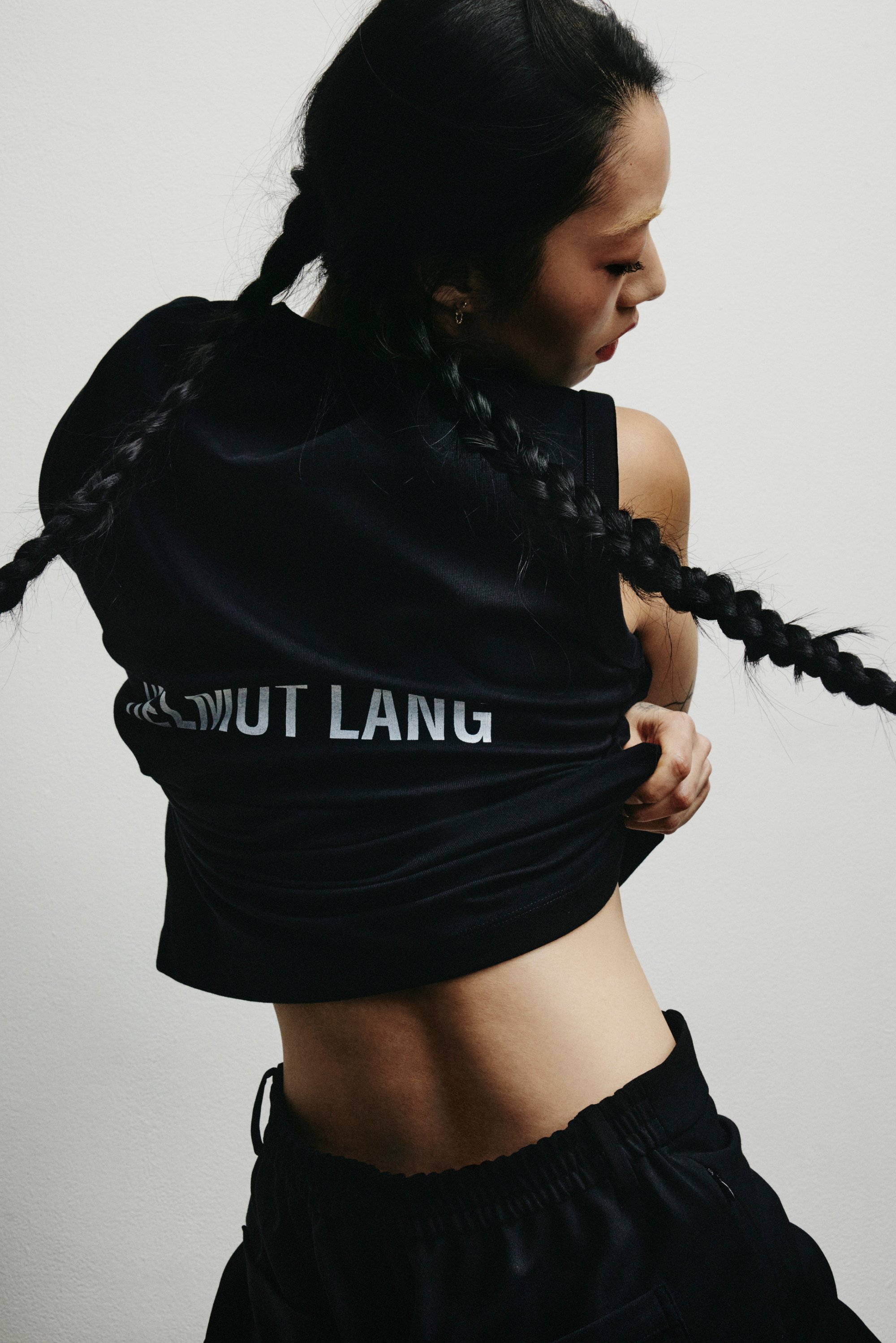 Una persona con largas trenzas levanta un top negro corto, revelando su abdomen. La parte posterior del top presenta el texto "NOT LANG."