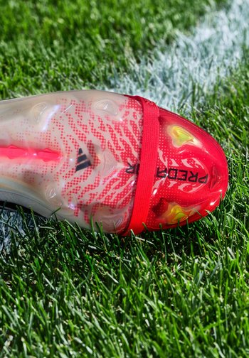 Chaussure de football rouge et blanche avec des crampons sur de l'herbe verte près d'une ligne blanche au sol. La chaussure porte la marque "Predator" avec un design texturé.