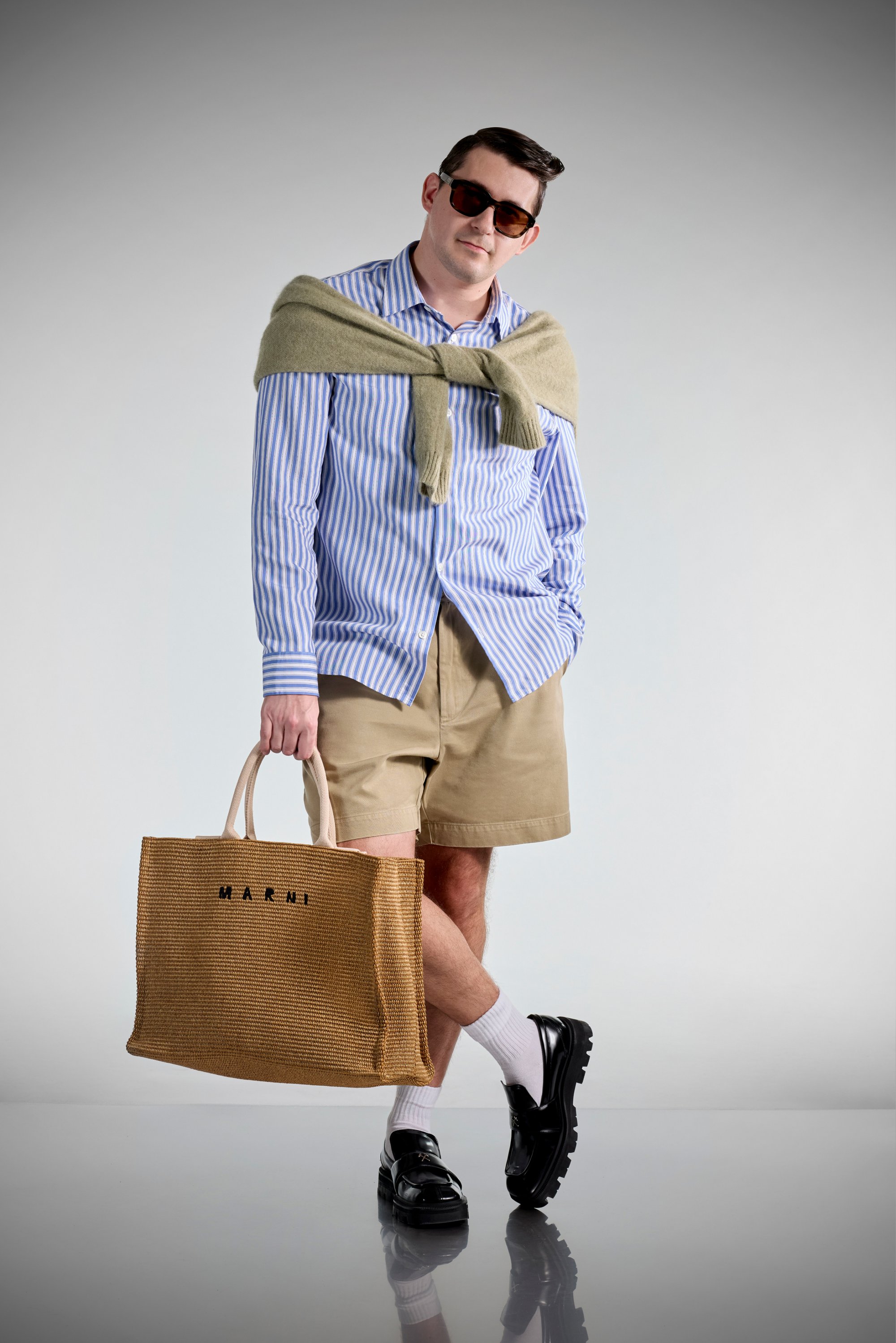 A man poses with a straw tote bag, wearing sunglasses, a striped shirt, khaki shorts, and black shoes, with a sweater draped over his shoulders.