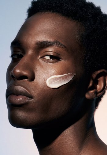 Jeune personne à la peau foncée et aux cheveux courts et bouclés avec une trace de crème blanche sur la joue droite, regardant légèrement de côté avec une expression neutre.