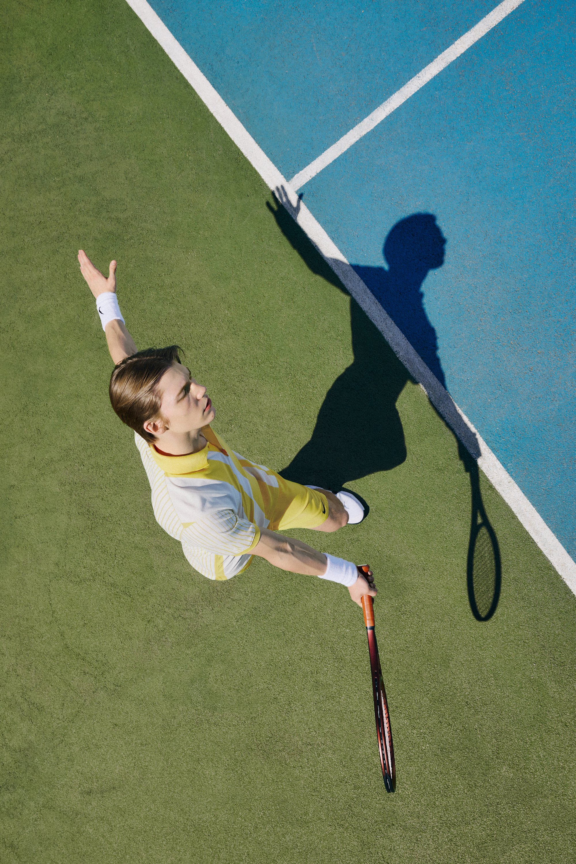 A young man in a yellow striped tennis outfit serves on a green court, his shadow stretching beside him, racquet held high.