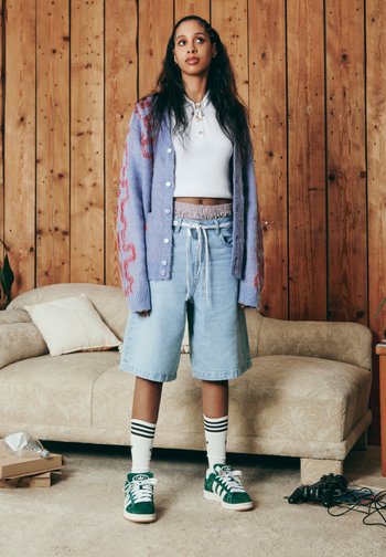 A young woman stands confidently in casual attire: a purple cardigan, white crop top, and denim shorts, in a cozy living room setting.