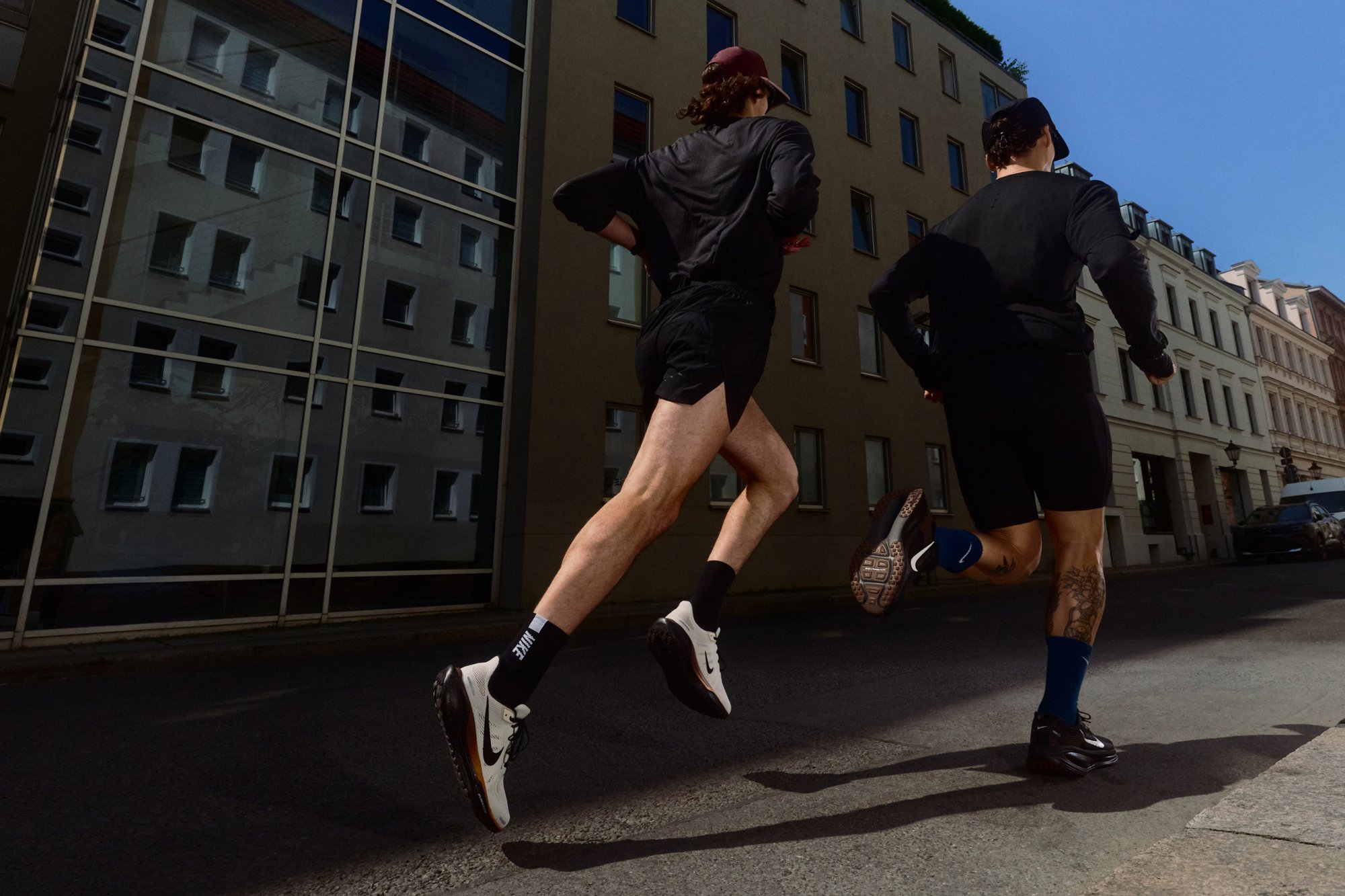 Two runners in black athletic shorts and tops jog along a city street. They are wearing black and white trainers, with one of them sporting blue socks.