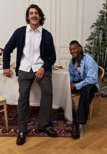 To menn sitter sammen i et rom. Den ene har på seg en marineblå cardigan over en hvit stripete skjorte, grå bukser og svarte sko. Den andre har på seg en lyseblå stripete skjorte og mørke jeans.