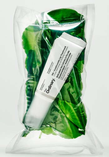 Un tube blanc de soin des lèvres The Ordinary HA repose en diagonale parmi des feuilles vertes, dans une pochette transparente scellée.