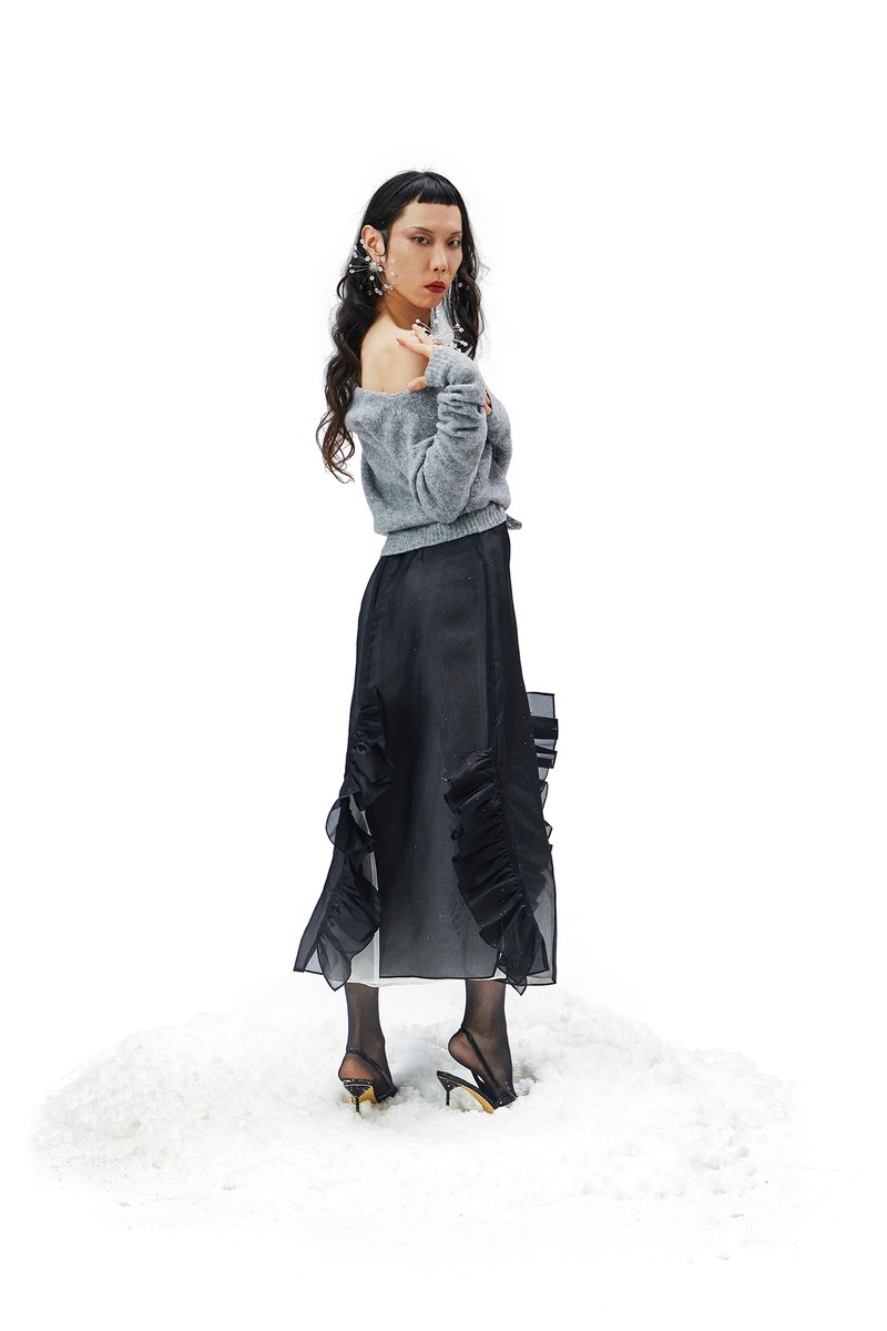 Una persona con lunghi capelli ondulati sta in piedi nella neve, indossando un maglione grigio scollo off-shoulder e una gonna nera a balze, posando con sicurezza.