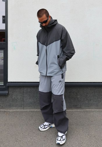 Casaco resistente ao tempo em cinza e cinza escuro com capuz e bolsos, combinado com calças a condizer. Os detalhes incluem o logótipo da Nike e um corte descontraído.