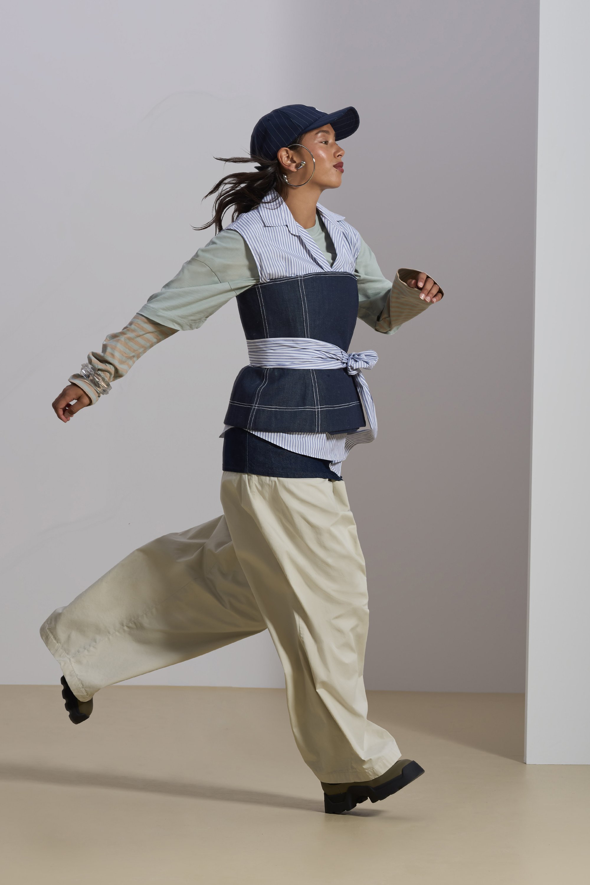 A person in a layered outfit, featuring a denim vest and wide light-colored pants, walks energetically against a simple background.