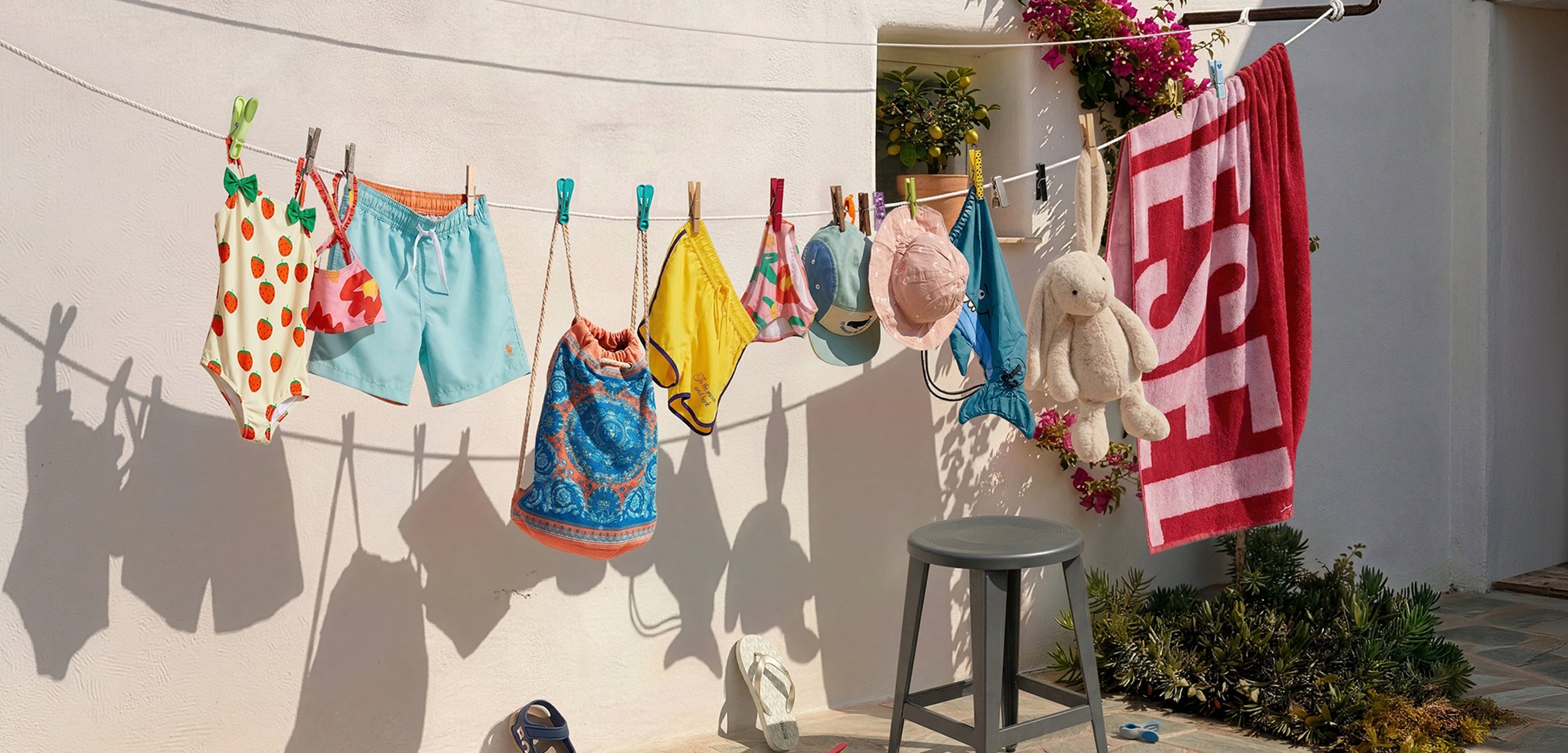 Ropa de baño de niños, sombreros, bolso, peluche y toalla colgados en un tendedero sobre una pared clara con sombras y un taburete gris cerca.