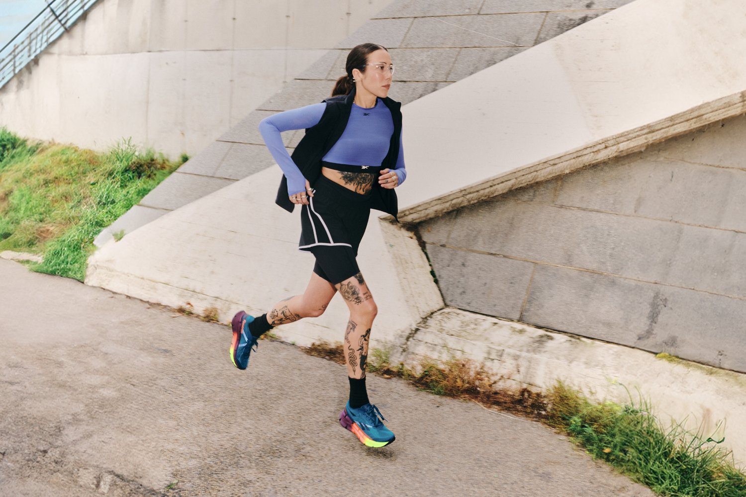 A tattooed woman runs on a path beside green grass, wearing a blue long-sleeve top, black shorts, and vibrant sneakers against a textured backdrop.
