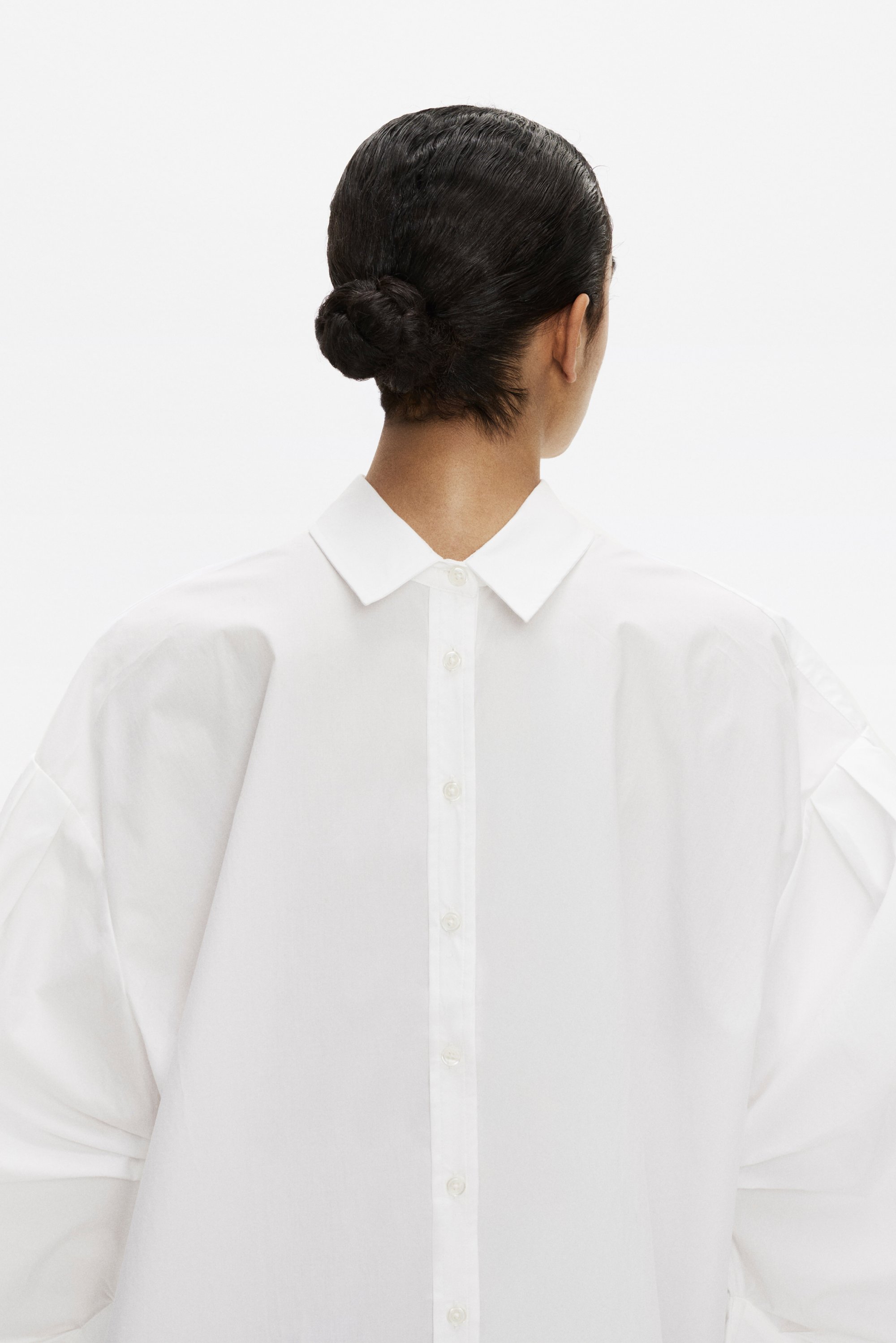 Una persona con un’acconciatura a bun è di spalle, indossa una camicia bianca oversize con un retro con bottoni e maniche larghe distintive.
