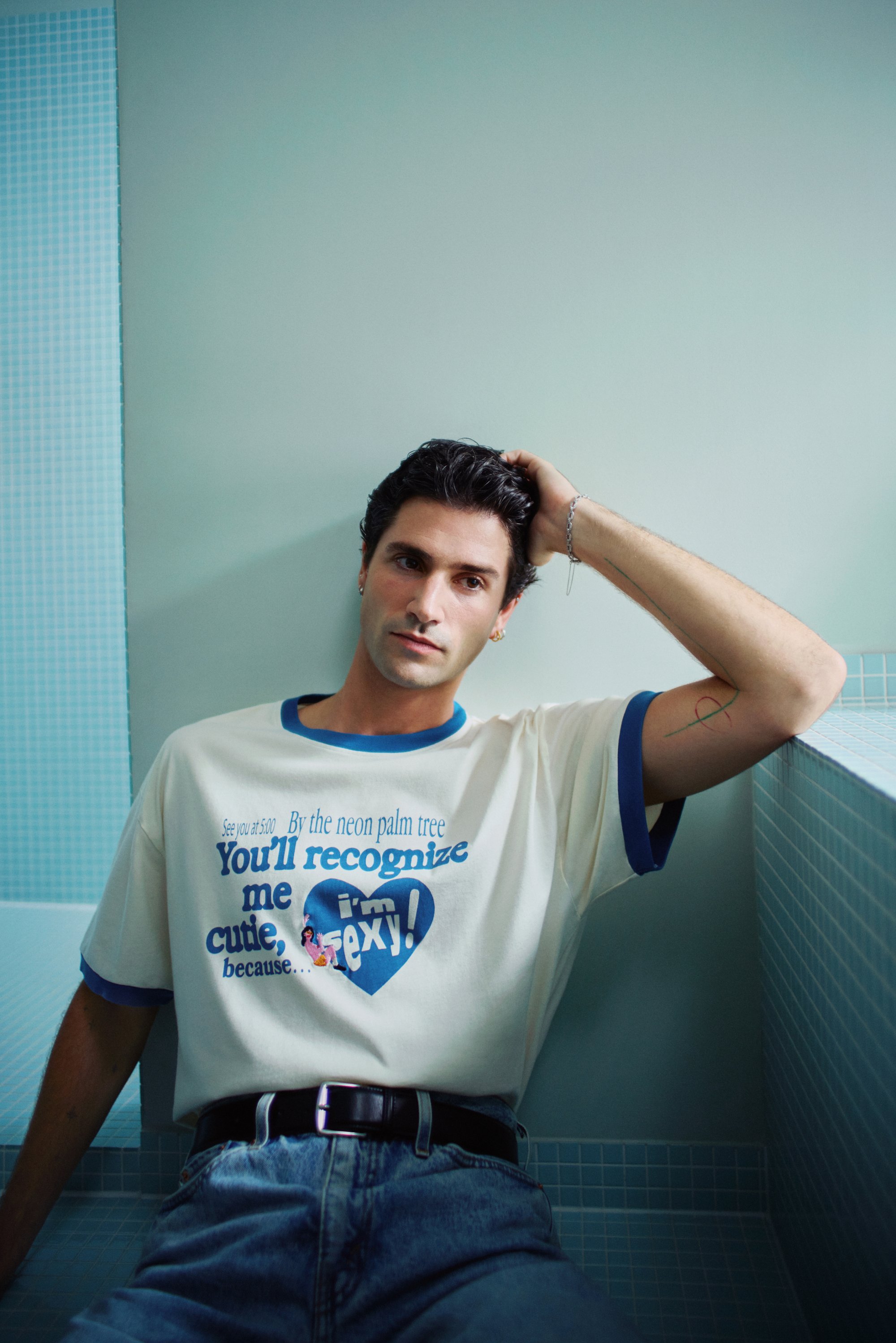 A young man with dark hair leans against a tiled wall, wearing a graphic t-shirt and jeans, looking thoughtfully at the camera.