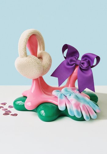 Pink abstrakter Ständer mit grünem Sockel, auf dem cremefarbene Ohrenschützer, eine lila Schleife und pastellblau-rosa Handschuhe gehalten werden, mit kleinem lila Herzkonfetti in der Nähe.