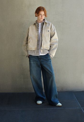 Casaco bege com forro texturizado, camisa cinza por baixo e jeans azuis de perna larga. Calçado é pálido e arredondado. Design simples e moderno.