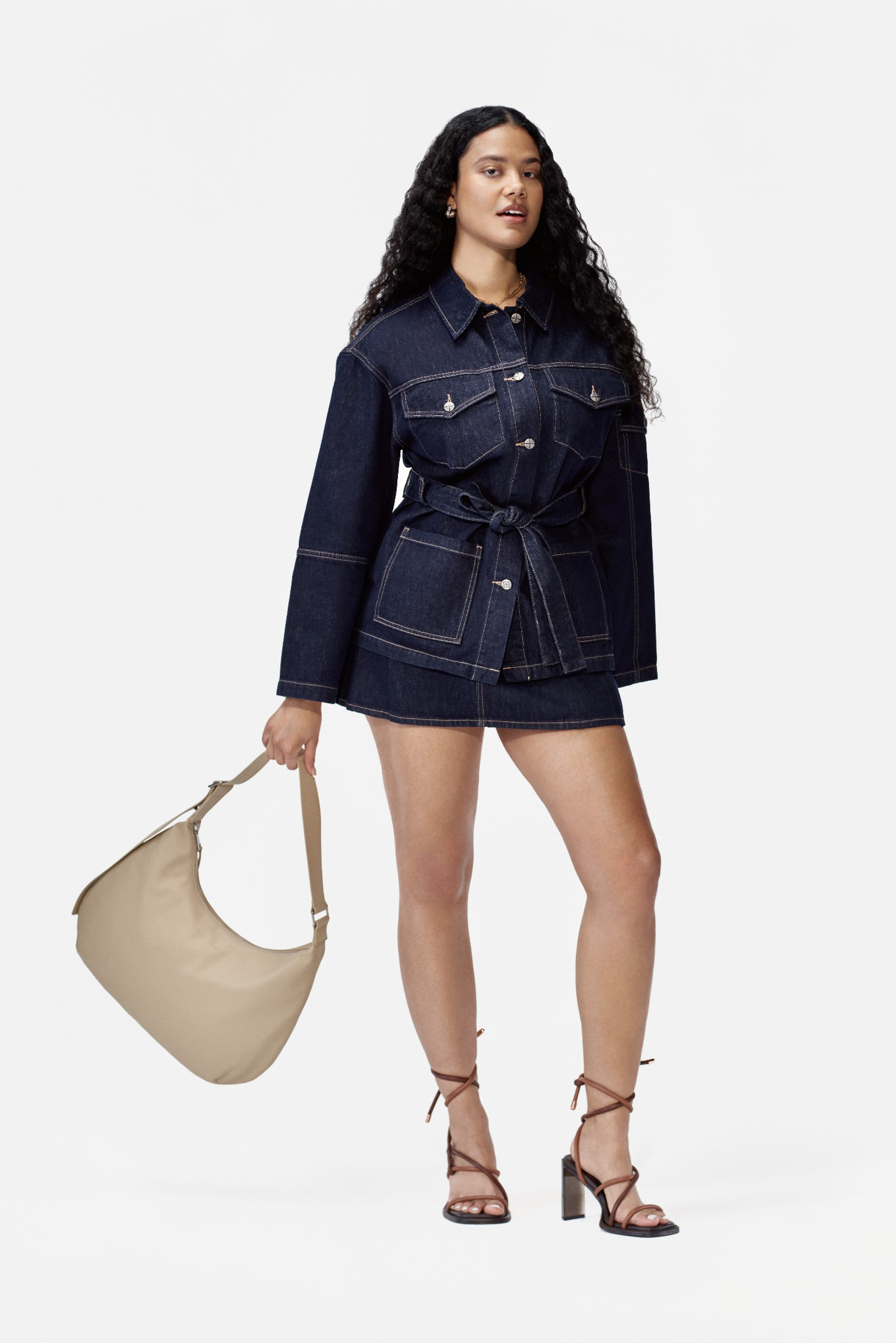 Una mujer con cabello rizado y largo lleva una chaqueta de mezclilla y una falda corta, sosteniendo un bolso beige, de pie contra un fondo blanco liso.