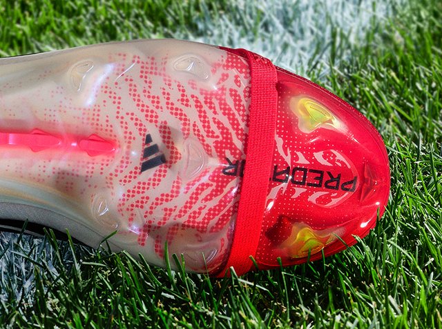 Közelről egy piros-fehér Adidas Predator focicipő fűben, megjelenítve a stoplikat és a texturált felsőrészt vörös pánttal.