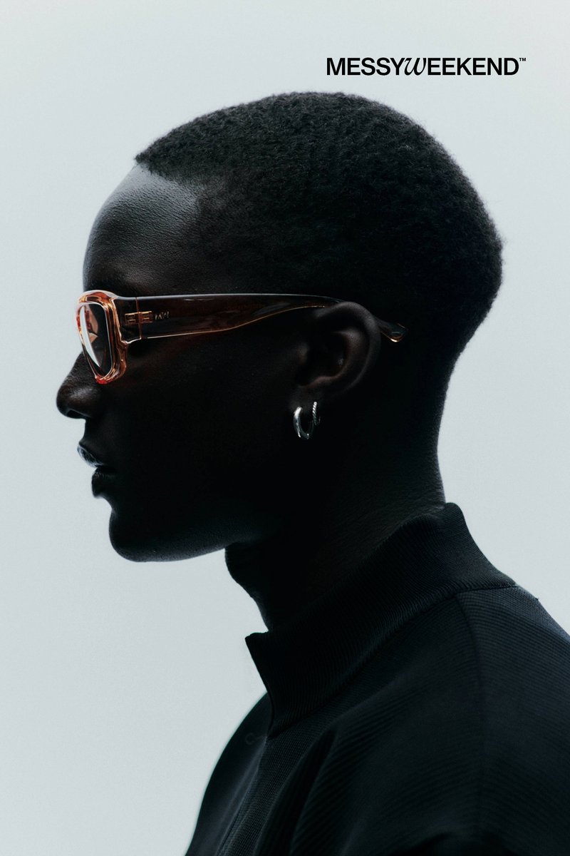 Perfil de persona con cabello corto y rizado que lleva gafas translúcidas marrones y pendiente de aro plateado sobre un fondo claro y liso.