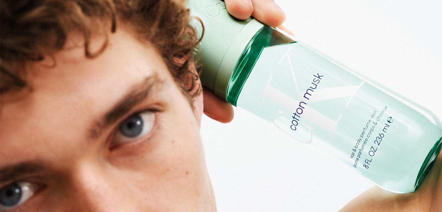 Man met krullend haar houdt een flesje met een groene dop en het etiket "cotton musk" dicht bij zijn gezicht tegen een witte achtergrond.
